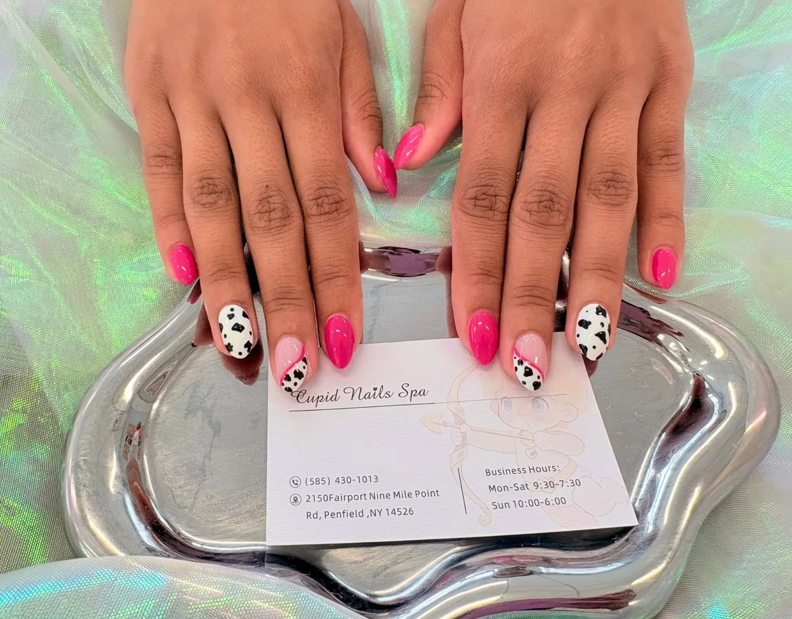 Hands with hot pink almond nails, two of them white with black cow-print spots and one a pink French tip over cow print, on a silver tray