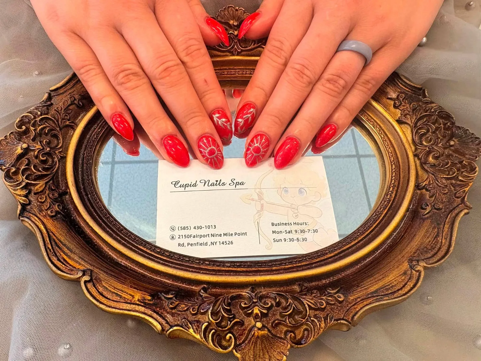 Two hands with long almond nails in glossy red, two of them painted with fine white sunbursts, resting on an ornate gold mirror